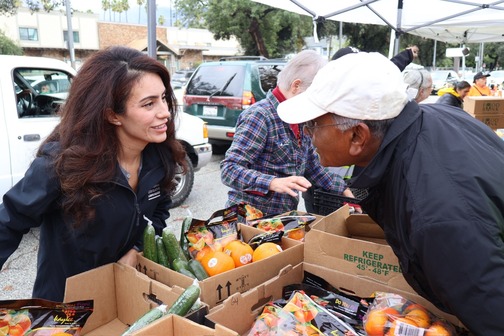 This image shows Senator Perez in a black jacket speaking with a man while standing at a table with boxes of food on it during her Operation Gobble turkey giveaway event. 