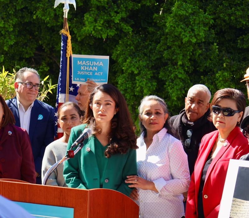 Wearing a green pantsuit, Senator Sasha Renee Perez stands at a podium and is surrounded by supporters of her bill, SB 995 the Masuma Khan Justice Act. 